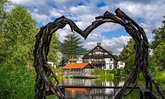 A picturesque house surrounded by greenery, seen through a heart-shaped wooden frame. In the foreground lies a tranquil pond that underscores the idyllic scene.