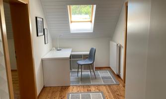 A bright room with a window and wooden floor. There is a desk with a chair and a lamp.