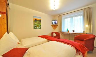 A cozy room with two beds and red accents. A picture of a landscape hangs on the wall, and there is a television as well as an armchair.