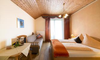 A cozy room with two beds and a wooden-paneled ceiling. There is a table, an armchair, and a terrace door with a view outside.