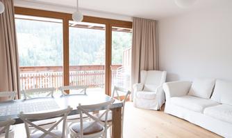 A bright and modern living room with large windows and a view of nature. There is a dining table and a comfortable couch.