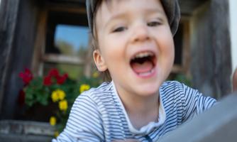 A laughing child with a hat stands in front of a window. Colorful flowers are blooming in the background.