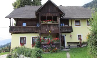 A cozy, yellow house with a wooden balcony and blooming flowers. Surrounded by green countryside and mountains.