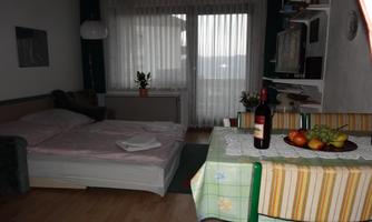 A cozy room with a double bed and a table set with fruit and a bottle of wine. Large windows let in plenty of light and offer a view outside.