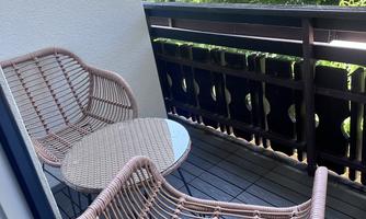 A cozy balcony with two rattan chairs and a small table. In the background, green trees are visible.