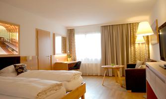 A modern hotel room with two beds and a large window. The decor is bright and inviting, with a table and a chair.