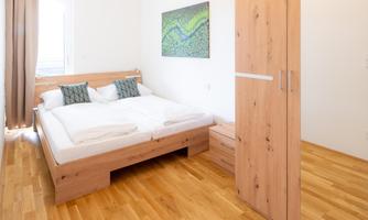 A bright bedroom with a double bed and two pillows. In the background, there is a wardrobe and a picturesque painting on the wall.