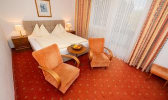 A cozy hotel room with a double bed and orange armchairs. The windows are adorned with curtains and plenty of daylight pours in.