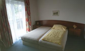 A simple bedroom with two single beds and a nightstand. The room has a large window with curtains and a picture on the wall.