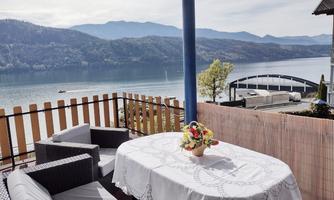 A terrace overlooking a tranquil lake and surrounding mountains. A flower decoration is on the table and there are cozy seating options available.