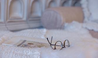 A stylish pair of glasses rests on a soft, white throw. In the background, an elegant, textured wall can be seen.