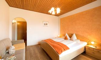 A cozy bedroom with a large double bed and a beige sofa. The walls are painted in warm tones, and there is soft lighting.