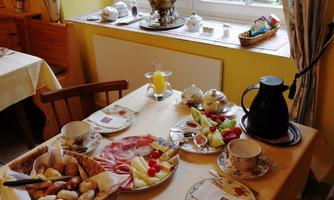 A nicely set table with a variety of breakfast items, some of them homemade and regional, including bread, cold cuts, and fresh fruit. A pot and cups are placed on a bright table in a welcoming room.