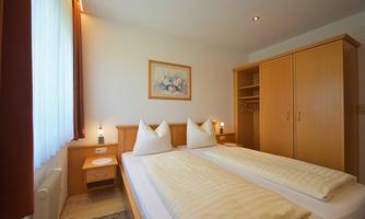 A cozy bedroom with two beds covered with white bedspreads. The walls are light, and there are wooden wardrobes as well as a window with curtains.