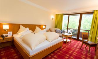 A cozy hotel room with a double bed, a sofa, and a table. Large windows offer a view of the green surroundings.