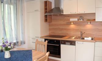 A modern kitchen with wooden and white furniture. A table with a flower vase and a blue tablecloth is visible.
