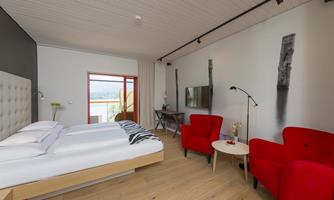 A modern hotel room with a large double bed and two red armchairs. The walls are bright, and there is a window with a view of the landscape.