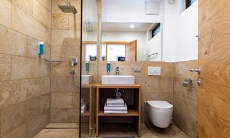 A modern bathroom with a glass shower and bright natural stone. It features a sink on a wooden shelf and a white toilet.