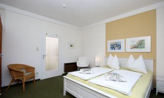 A bright bedroom with a double bed and two pillows. There is a chair and a rattan chair, as well as some artworks on the wall.