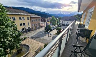 A view from a balcony overlooking a quiet city street with colorful houses. In the background, mountains and a cloudy sky can be seen.