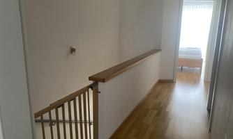 A bright stairwell with a wooden floor and a railing. At the end of the corridor, a window can be seen, letting in natural light.