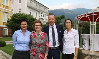A group of four people is smiling warmly in a garden with green surroundings. Buildings and mountains can be seen in the background.
