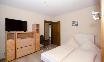 A simple bedroom with a double bed, a television, and wooden cabinets. The walls are painted in a neutral color and there is a chair as well as a picture on the wall.