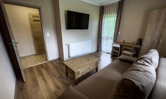 A cozy living space with a sofa, a table, and a TV on the wall. The bright walls and the wooden floor create an inviting atmosphere.