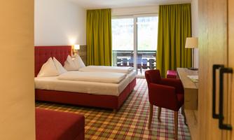 A modern hotel room with two beds, a desk, and a balcony. The walls are light, and the curtains are in a vibrant green.