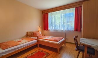 A simple room with two single beds, a table, and a chair. The room has large windows with bright curtains and warm colors.