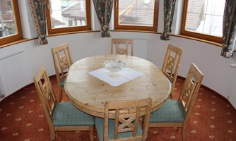 A round wooden table with six chairs in a bright room. The windows are adorned with curtains and the floor is covered with a carpet.