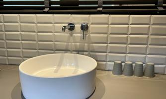 A classic, slightly rounded sink on a modern shelf. In the background, there is a textured wall and a few small cups.