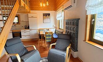 A cozy room with wooden paneling and large windows. Two comfortable armchairs are positioned on a carpet, while a modern kitchen is visible in the background.