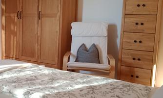 A cozy bedroom with a wooden wardrobe and a chest of drawers. A decorative blanket is on the bed and a comfortable armchair stands next to the bed.