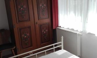 A cozy bedroom with a white bed and a decorative wardrobe. The light gently falls through the curtains, creating an inviting atmosphere.