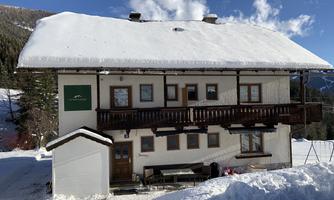 A cozy house in the snow with a balcony and an inviting front door. The surrounding landscape is characterized by mountains and pine trees.