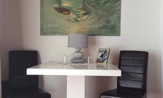 A modern table with two black chairs is in a room. A painting depicting a coastal scene with a boat is hanging on the wall.