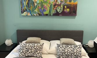 A modern bedroom with a gray bed and striking cushions. Above the bed hangs a colorful piece of artwork featuring faces and plant motifs.