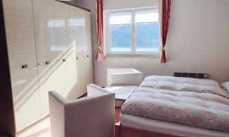 A simple bedroom with a large bed and two white armchairs. The room has light walls and a window that lets in daylight.