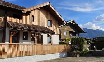 A modern wooden house with a balcony and a beautiful terrace. In the background, there are mountains and a clear sky.