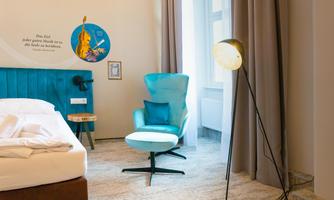 A modern hotel room with a cozy bed and a blue armchair. The room has large windows with curtains and a stylish floor lamp.