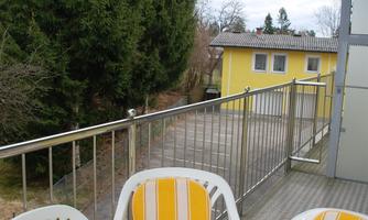 A balcony with two chairs and a round table. In the background, a yellow house and a large tree can be seen.