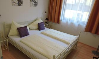 A cozy bedroom with a double bed and purple pillows. The windows are covered with white curtains and there are decorative pictures on the wall.