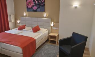 A cozy hotel room with a double bed and a black armchair. The walls are painted in warm colors, and a floral painting adorns the wall above the bed.