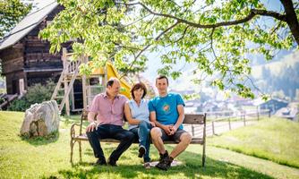 Three people are sitting relaxed on a bench under a tree. In the background, a wooden building and a green landscape can be seen.
