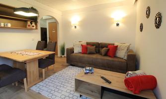 A cozy living room with a brown sofa, red cushions, and a wooden table. The walls are light, and there is friendly lighting.