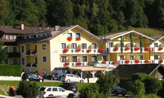 A cozy hotel with a yellow facade and balcony flowers. Surrounded by green trees and parked cars.