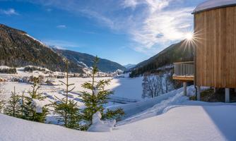 A wintry landscape with snow-covered mountains and a sunny sky. In the foreground, there are some fir trees and a modern wooden building.