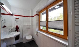 A modern bathroom with white tiles and a window that lets in daylight. There is a toilet, a sink, and towels in red colors.