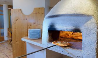 A large wood oven in a restaurant, from which a freshly baked pizza is being taken out. Stacked plates and wooden walls are visible in the background.